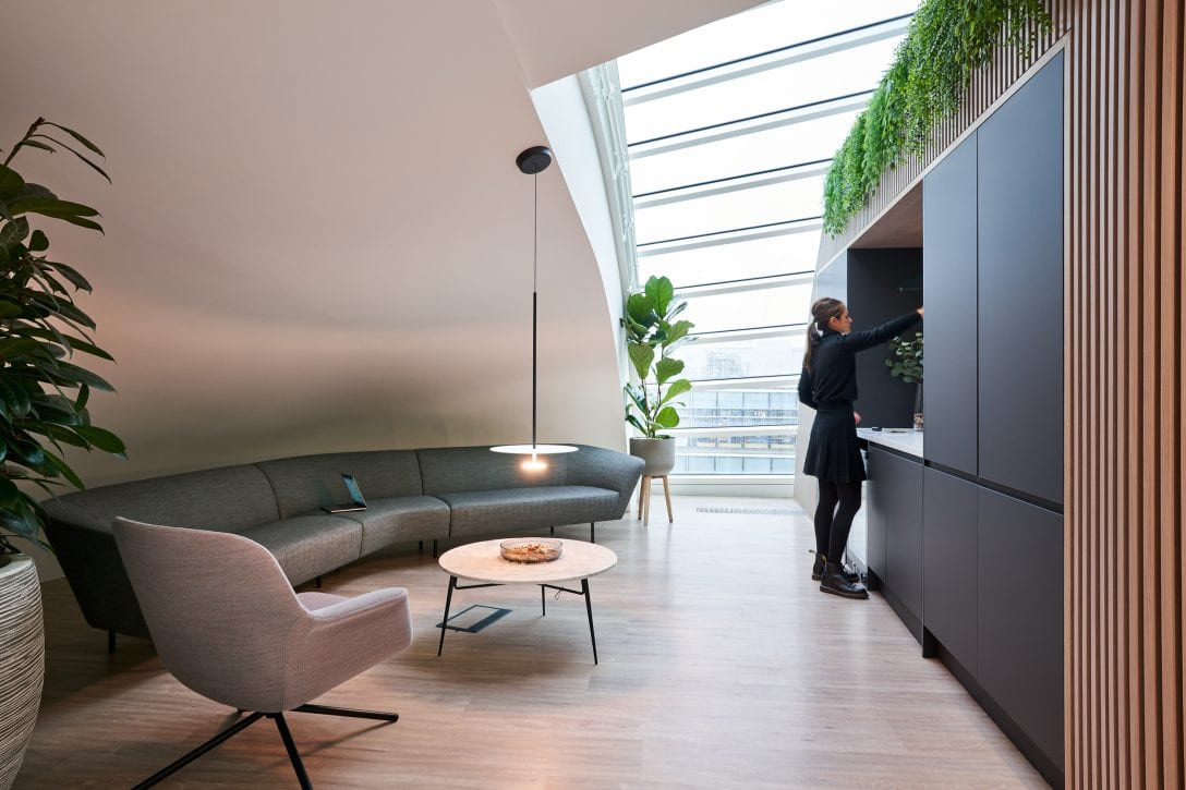 A woman is shown inside an office reaching for a class in a tea point. Behind her is a sofa with an open laptop resting on it. 