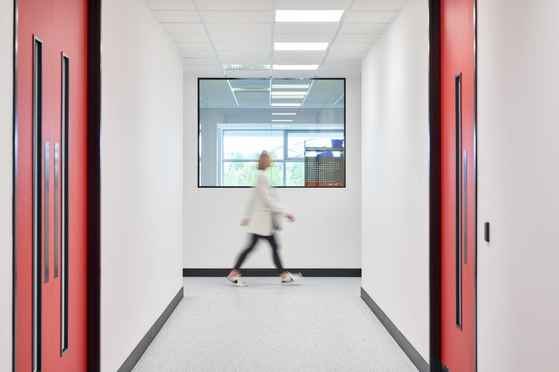Female lab professional in white lab coat walks past the window of an enclosed laboratory space.
