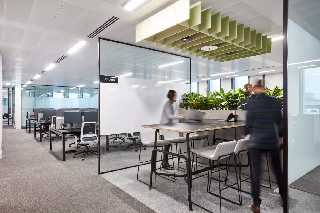 Two employees are shown about to sit down at a high collaboration bench inside an office. 